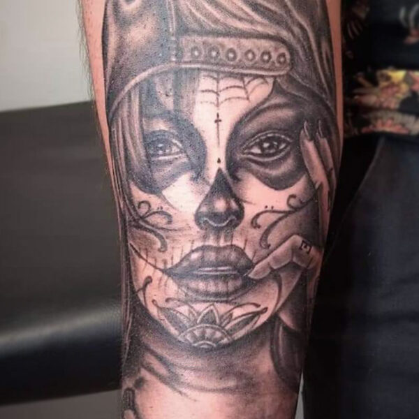 atticus tattoo, black and grey realism tattoo of a woman with a sugar skull face and wearing a backwards cap