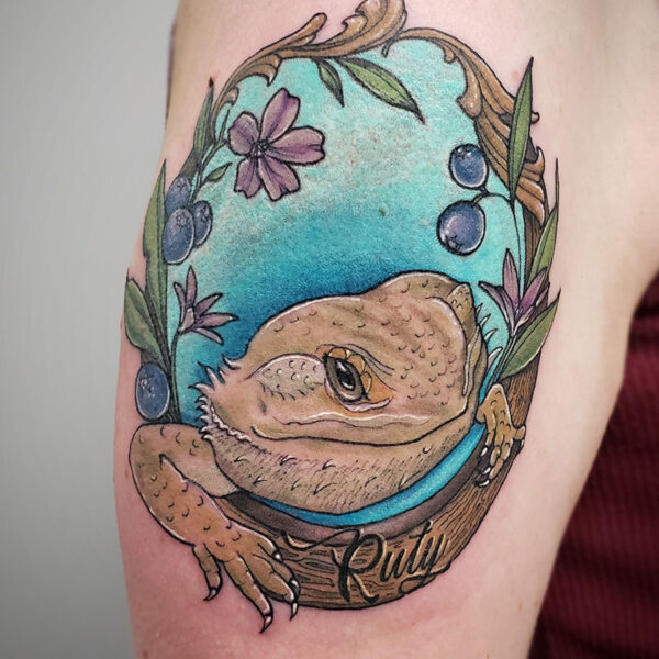 atticus tattoo, neotraditional tattoo of a bearded dragon in frame with flowers, berries and a blue background