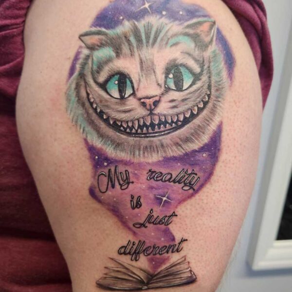 atticus tattoo, coloured tattoo of the Cheshire Cat coming out if a book with the words "My reality is just different"