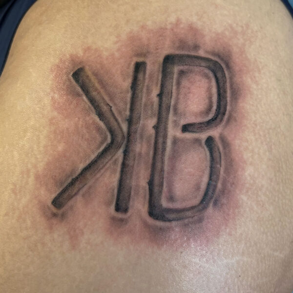 atticus tattoo; black and grey tattoo of a brand of a backwards "k" and the letter "b"