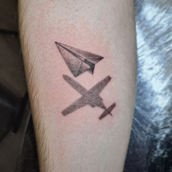 atticus tattoo, black and grey tattoo of a paper plane with the shadow of a fighter plane