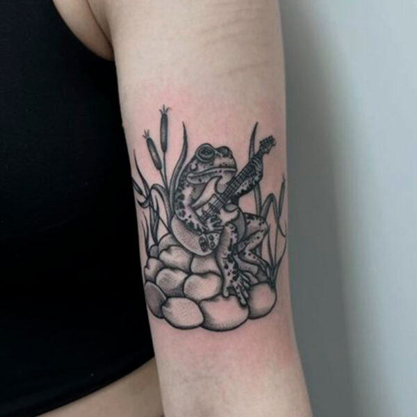 atticus tattoo, black and grey tattoo of a frog sitting on rocks while playing a guitar