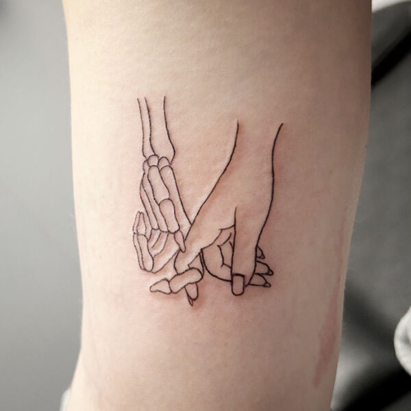 atticus tattoo, line tattoo of a skeleton hand and woman's hand holding each others fingers