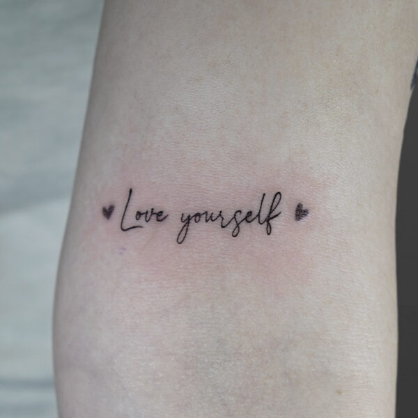 Atticus Tattoo| Fine line tattoo of the words "Love Yourself" with a heart on each side