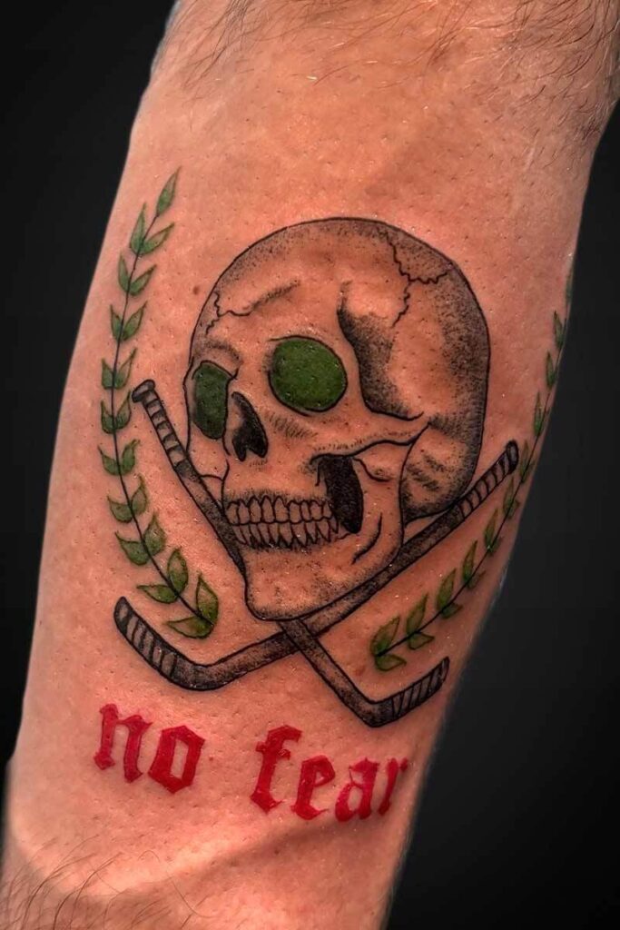 Atticus Tattoo| Black and grey tattoo of a skull with hockey sticks behind it, green leaves and red script that says "No fear"