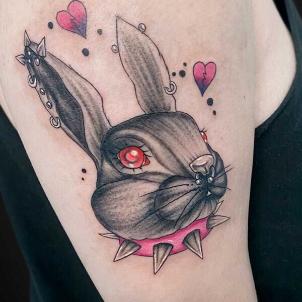 Close-up of a bunny tattoo: gray-black shading, red eye, pierced ears, pink spiked collar, and floating pink hearts on the upper arm.