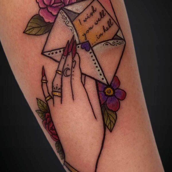 Tattoo of two hands with long nails holding a geometric origami envelope, pink flowers, and leaves, with handwritten words “I wish you were here.”