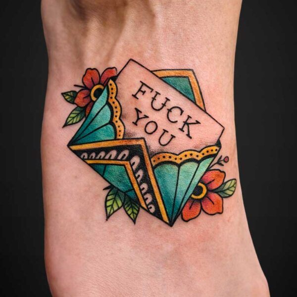 Colorful traditional-style tattoo on the ankle featuring playing cards with the bold phrase 'FUCK YOU' and orange/red flowers nearby.