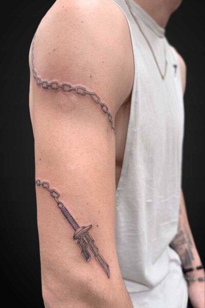 Arm tattoo featuring a chain wrapping from shoulder to mid-upper arm and a dagger tattoo on the forearm area.