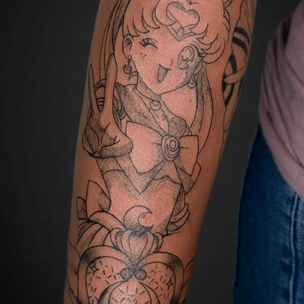 Line-drawn Sailor Moon tattoo on a forearm, shows her winking and making a peace/gesture with a heart motif and floral accents below