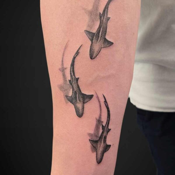 Forearm tattoo of three stylized black fish swimming upward with flowing tails on the skin.