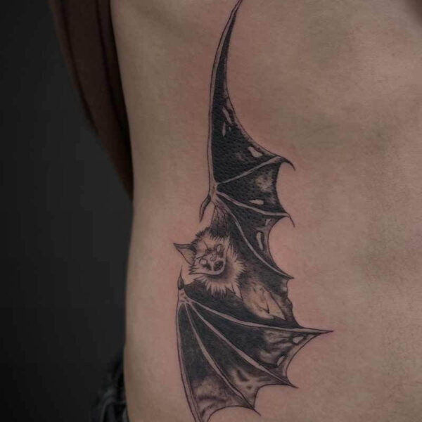 Tattoo of a bat with a cat-like face on the side of the torso, black shaded wings wrapping down the rib area.