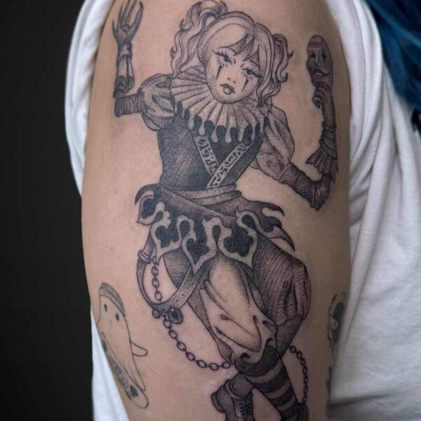 Black-and-gray tattoo of a jester/clown woman with a ruffled collar, raised arms, and decorative armor on the upper arm.
