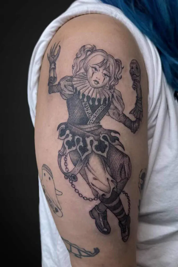 Black-and-gray tattoo of a jester/clown woman with a ruffled collar, raised arms, and decorative armor on the upper arm.