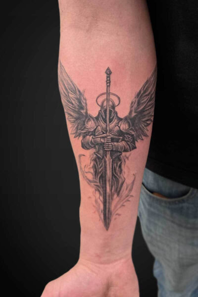 Detailed black-and-gray tattoo of a winged armored knight gripping a long sword along the forearm, sword pointing downward.