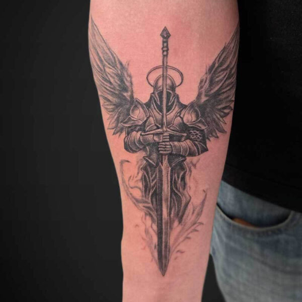 Detailed black-and-gray tattoo of a winged armored knight gripping a long sword along the forearm, sword pointing downward.