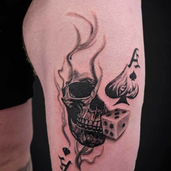 Tattoo on the upper arm of a skull with flames and smoke, a dice, and spade symbols in black and gray ink.