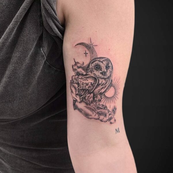 Tattoo on the upper arm of an owl perched on rocks with a crescent moon, sunburst, and stars, plus a small 'M' below.