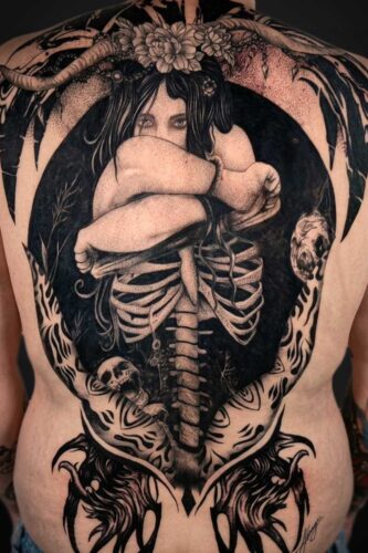 Back tattoo showing a curled woman inside a dark circular frame, with an exposed ribcage and skulls around the edges.