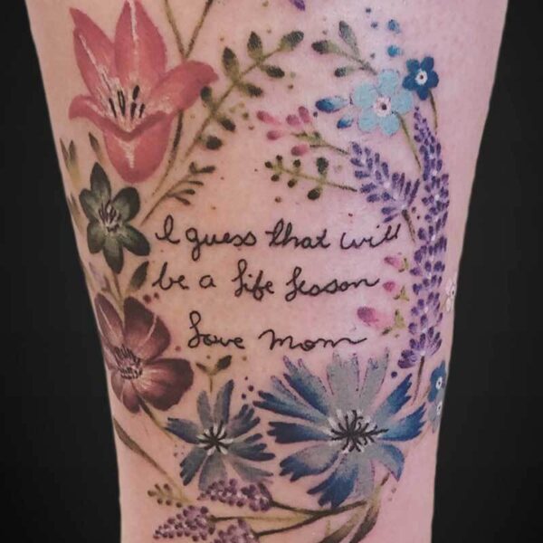 Colorful floral tattoo surrounding a handwritten quote: 'I guess that will be a life lesson — Love Mom' on the calf.