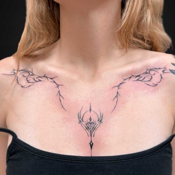 Close-up of a woman's décolletage with intricate thorny tribal tattoos along the collarbones and a central ornate design at the sternum, wearing a black top.
