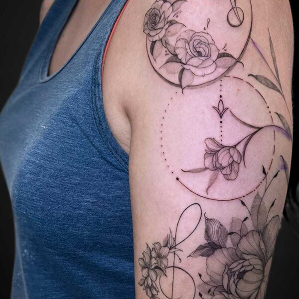 Upper-arm tattoo with roses and delicate linework, including circular and dotted geometric elements on a floral sleeve.