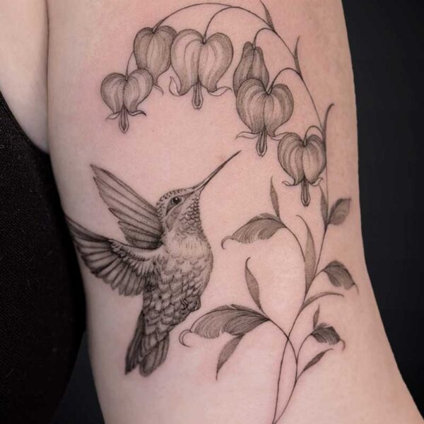 Delicate black‑gray tattoo of a hummingbird hovering beside a curved stem with heart-shaped bleeding‑heart blossoms on the upper arm.