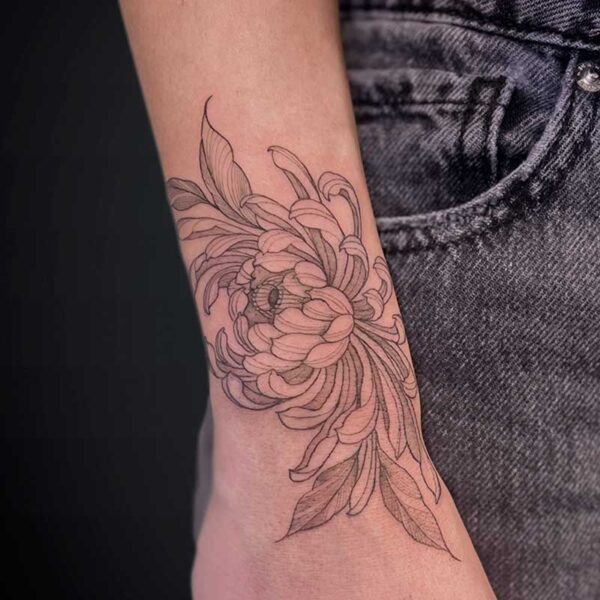 Line-drawing floral tattoo wrapping around the forearm near the wrist, with detailed petals and leaves, plus a small script tattoo on the underside of the wrist.