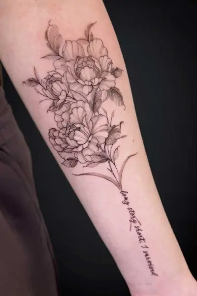Black linework floral tattoo along the forearm featuring blooming peonies and leaves, with a cursive inscription along the lower arm. The handwritten text is present but not legible in the image.