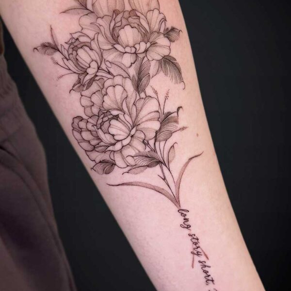 Black linework floral tattoo along the forearm featuring blooming peonies and leaves, with a cursive inscription along the lower arm. The handwritten text is present but not legible in the image.