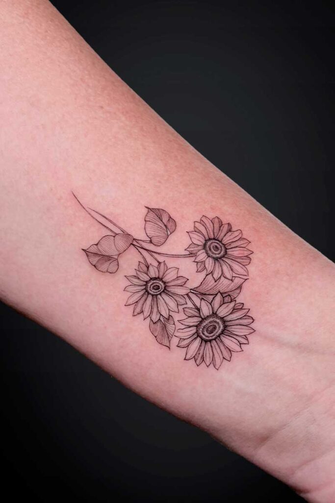 Line-drawn tattoo of three sunflowers with leaves on the inner ankle.
