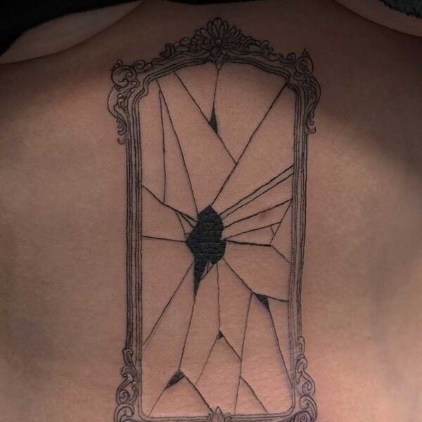 Ornate vertical rectangular frame tattoo on skin showing a shattered mirror with jagged shards and a dark central break point.