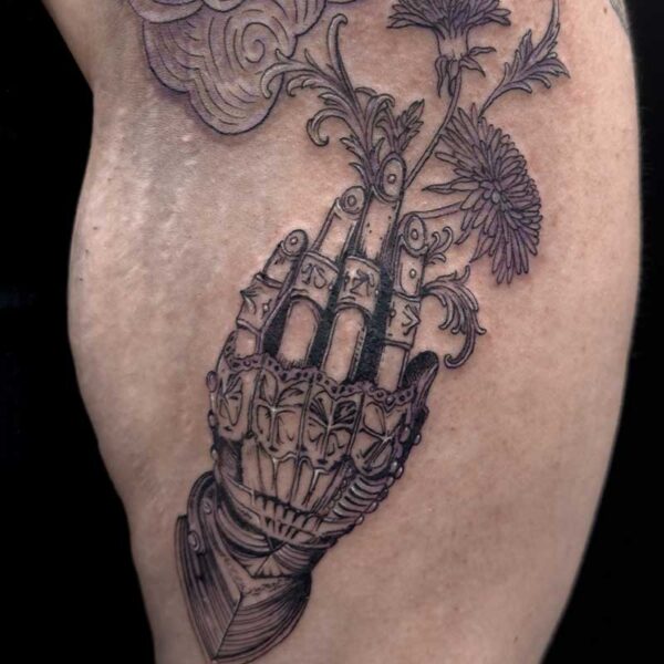 Tattooed forearm featuring a detailed mechanical-looking hand with metal segments, from which wildflowers grow, with stylized clouds above.