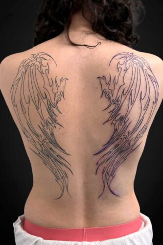 Back view of a person with large black wing outlines tattooed across the shoulder blades on an exposed back.