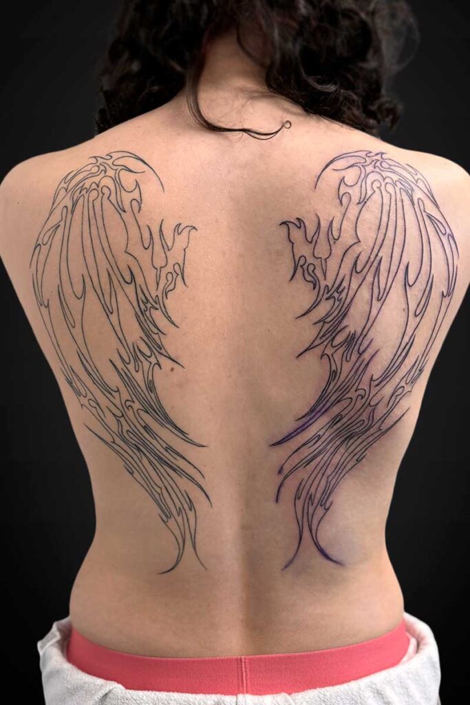 Back view of a person with large black wing outlines tattooed across the shoulder blades on an exposed back.