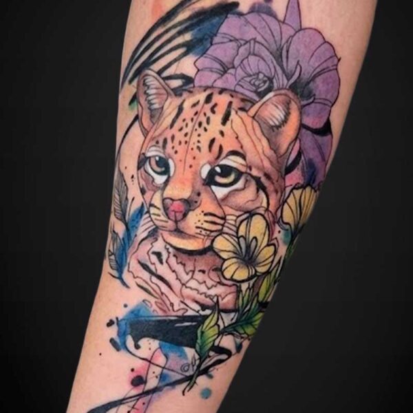 Colorful watercolor-style tattoo of a leopard cub surrounded by purple and yellow flowers on a forearm.