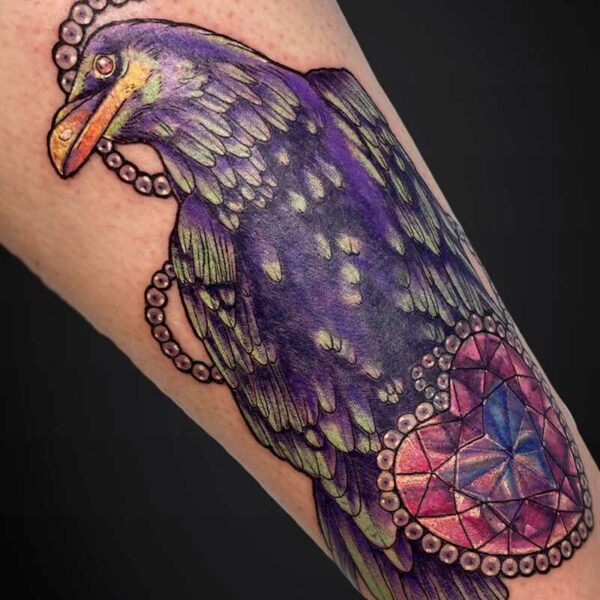 Colorful raven tattoo on the forearm with purple feathers and green accents, encircled by a beaded chain and a pink geometric gem here-and-there on the arm.