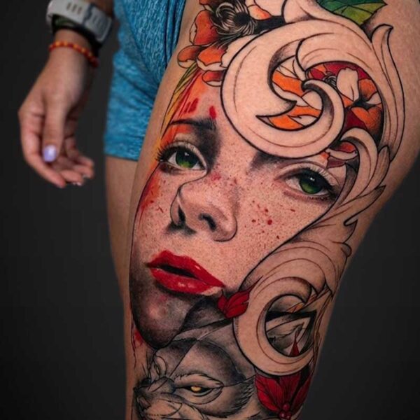 Colorful tattoo on a thigh featuring a woman's face with green eyes and red lips, surrounded by flowers and ornamental swirls, with a wolf-like animal at the lower edge.