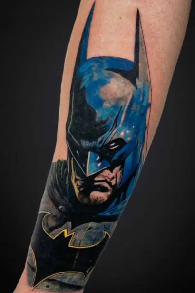 Close-up of a Batman cape and face tattoo on a forearm, blue and black ink with yellow accents forming the Batman emblem at the wrist area