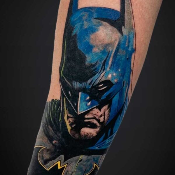 Close-up of a Batman cape and face tattoo on a forearm, blue and black ink with yellow accents forming the Batman emblem at the wrist area