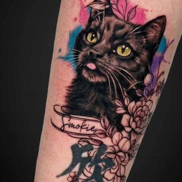 Tattoo of a black cat with yellow eyes and a pink tongue, surrounded by watercolor blue and pink splashes and floral line art, with a ribbon reading Smokie.