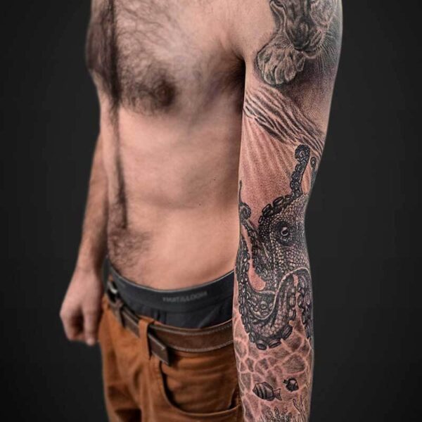 Side view of a shirtless man with a detailed black ink sleeve tattoo on his right arm, featuring an octopus and underwater motifs, wearing brown cargo shorts.