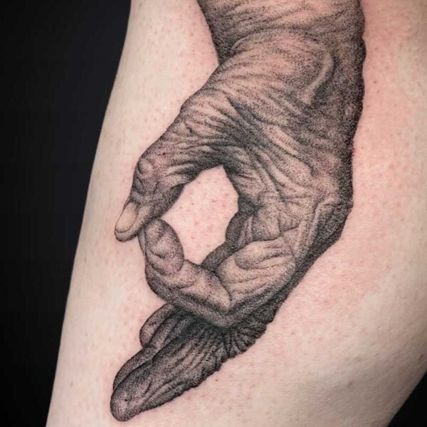 Realistic black-and-gray tattoo of a hand forming an OK gesture with detailed wrinkles, on the upper arm, with a feathered extension beneath the wrist.