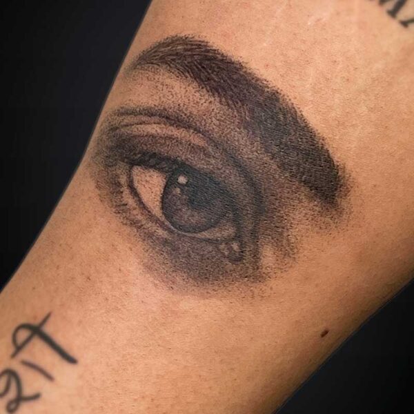Close-up of a hyper-realistic black-and-gray eye tattoo on a forearm with detailed shading around the lid and iris.