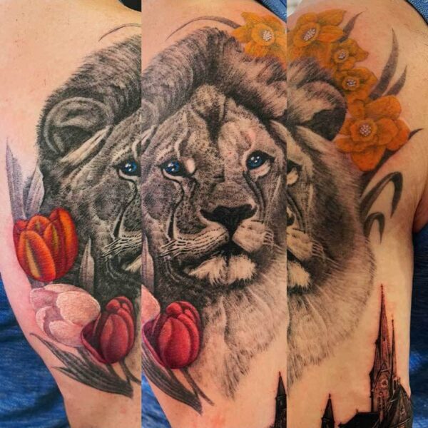 Realistic lion portrait tattoo on the upper arm, with orange flowers on the right and red tulips on the left, plus a partial skyline at the bottom arc of the design.