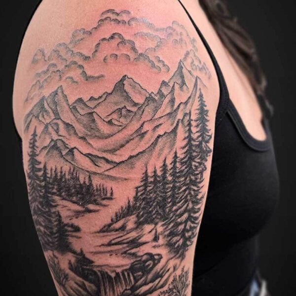 Black-and-gray mountain landscape tattoo wrapping the upper arm, featuring jagged peaks, evergreen trees, and a flowing stream or small waterfall under a cloudy sky.