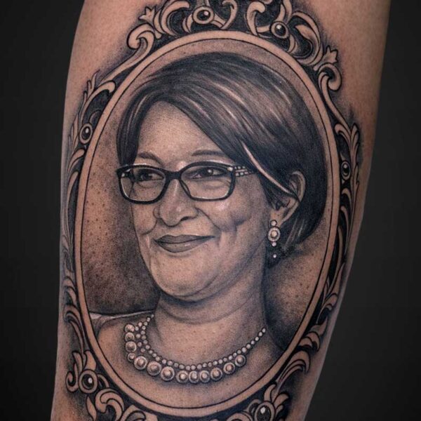 Tattoo of an older woman with short dark hair, glasses, and a pearl necklace in an ornate oval frame.
