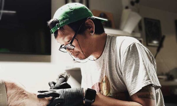 Tattoo artist in a green cap and glasses tattoos a client's leg on a padded table, wearing gloves and a smartwatch in a studio.