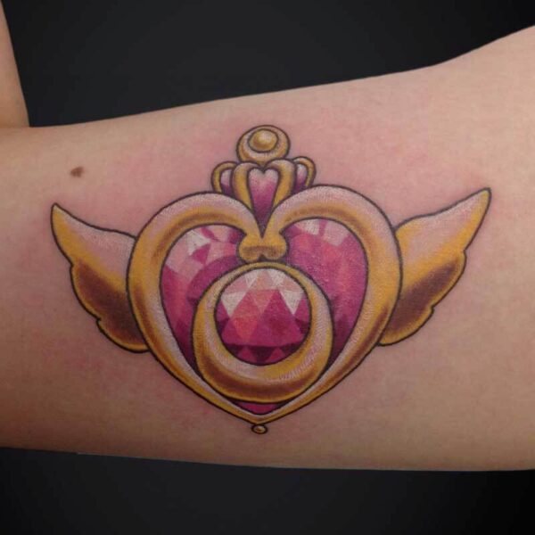 Tattoo of a pink gem heart with a gold ornamental frame and small wings on the forearm.