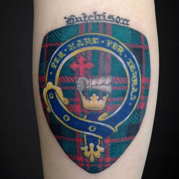 Tattoo of a heraldic shield on a plaid background, featuring a gold crown, blue ribbon, red cross, and a surrounding yellow ring, with a scripted word above the shield.